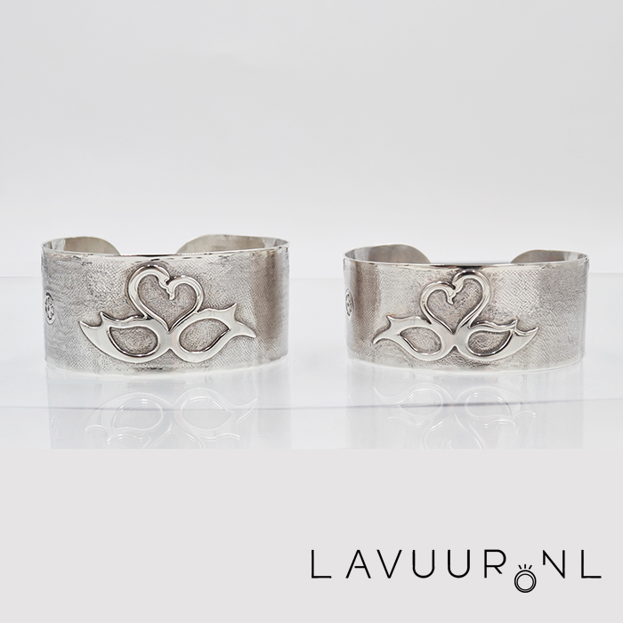 twee zilveren armbanden van breed plaat, ruw gestempeld en elk voorzien van een opengewerkt figuur van twee zwanen in een hart er op gesoldeerd